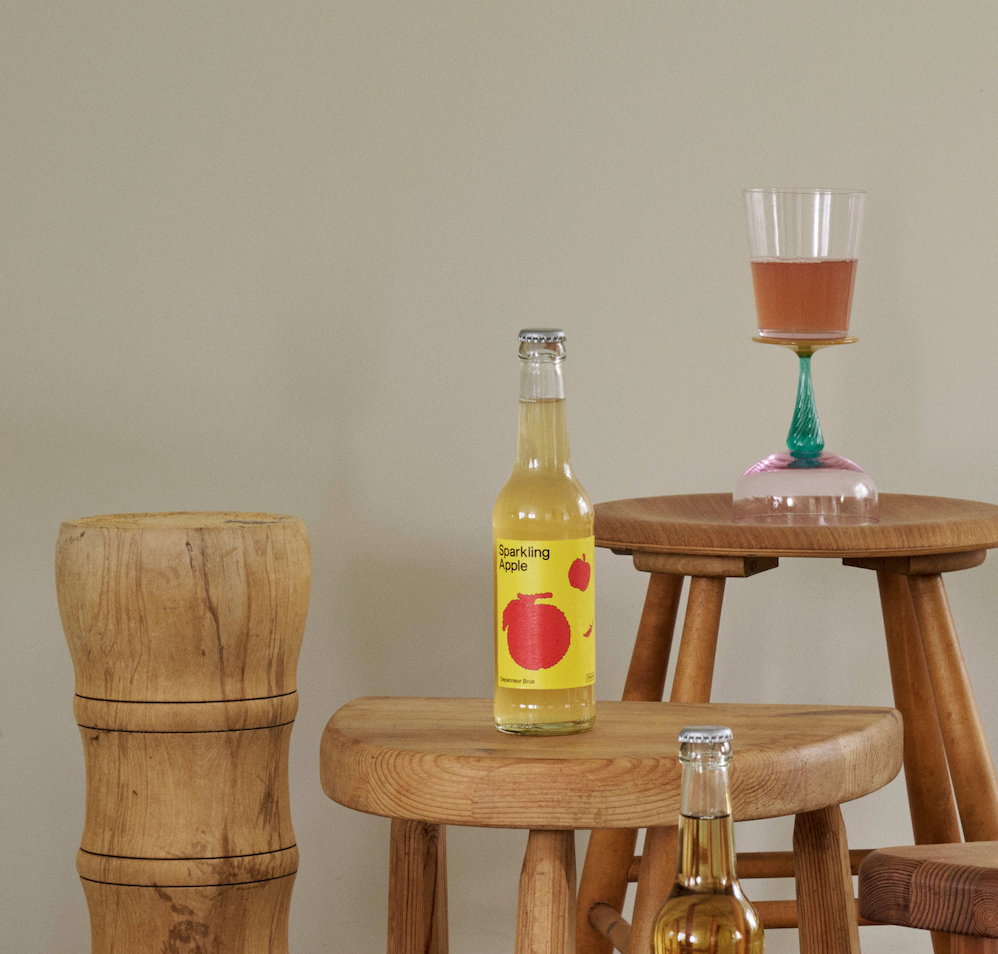 Bottle of Depanneur BRUS Sparkling Apple juice on a wooden stool with a glass of juice in the background.