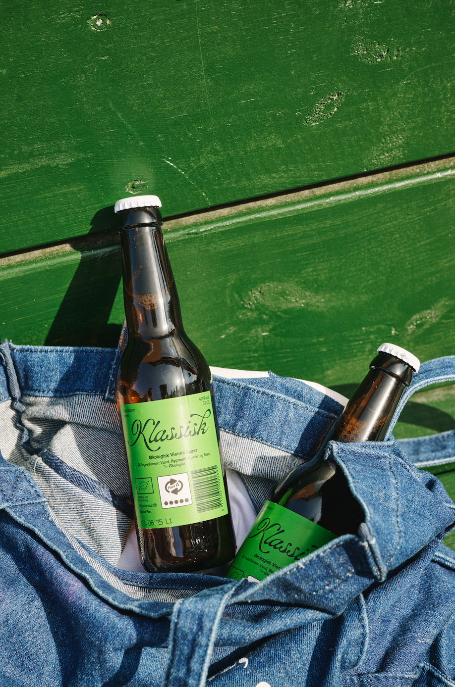 Two bottles of Klassik beer with green labels in a blue denim jacket against a green background.
