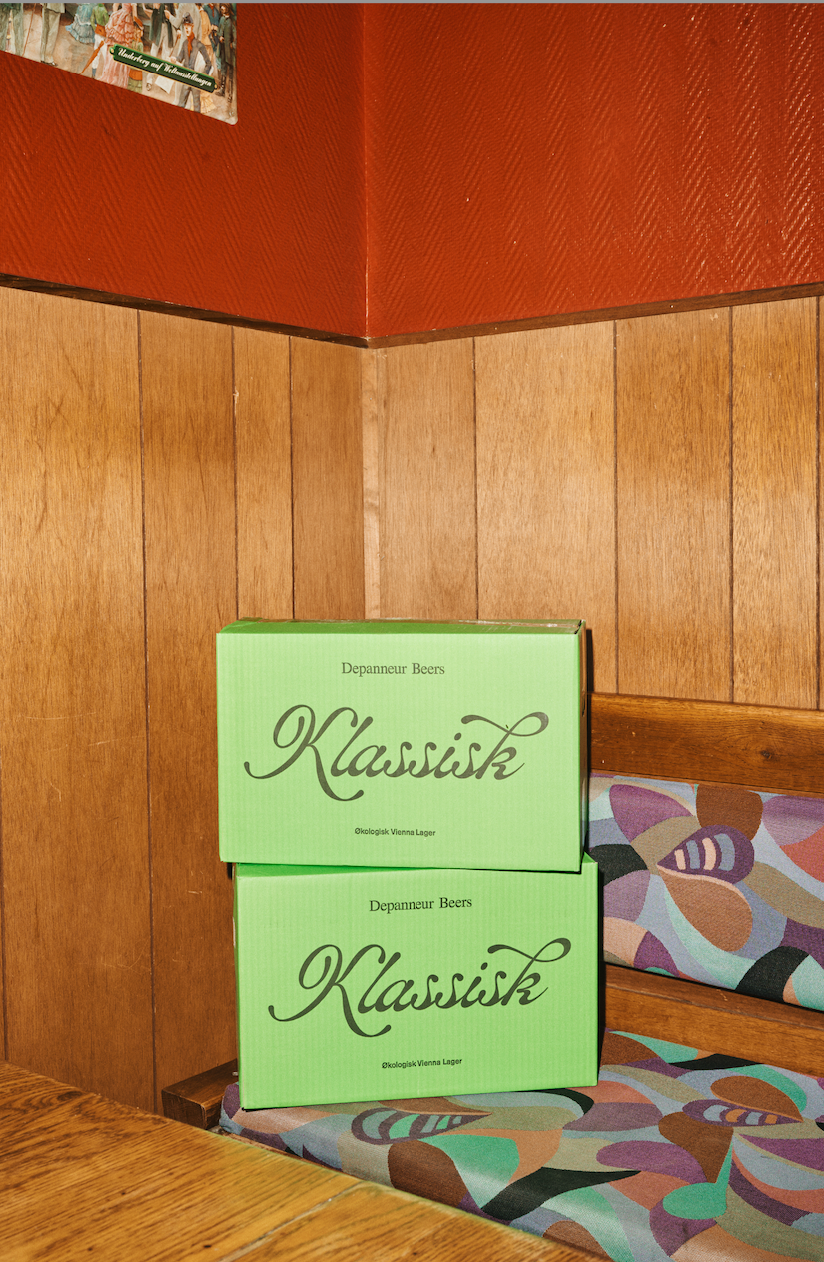 Two green organic cases labeled 'Klassisk' stacked on a wooden surface with a patterned cushion in the background.